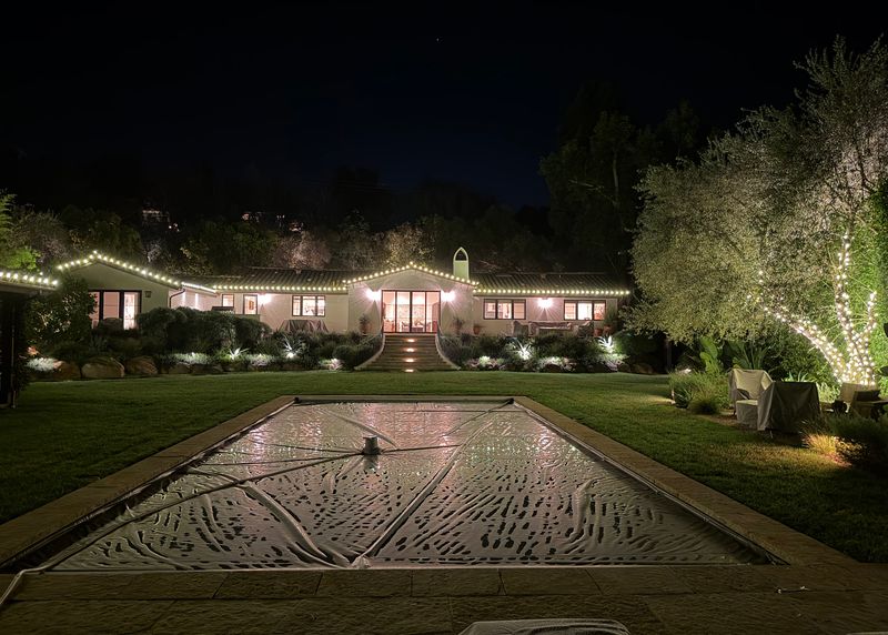 After: the same home at night with warm-white lights along the roofline and a lit olive tree