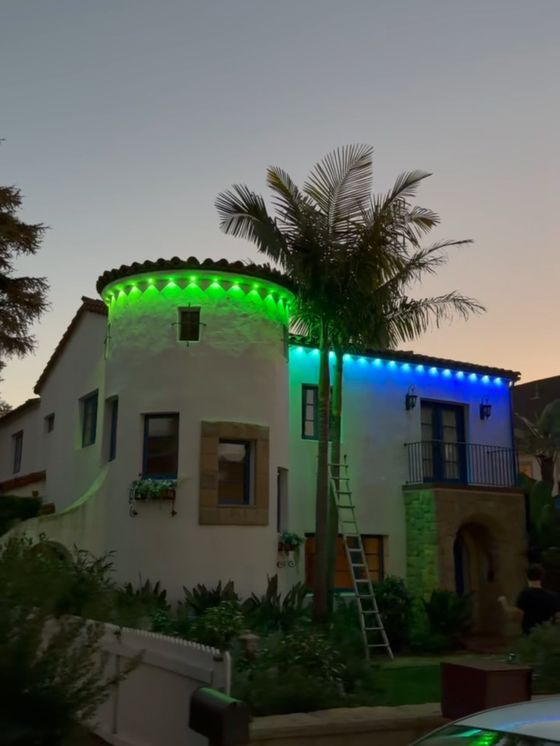 A Spanish-style home at dusk with permanent color-changing lights along the roofline, glowing green and blue